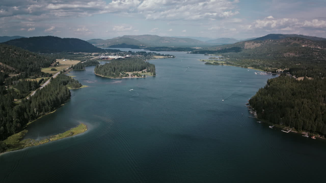 Aerial timelapse video of Priest River flowing past forested islands and peninsulas, with speed boats in blue water reflecting the sky and mountains rising in the distance