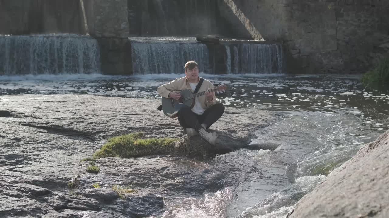 바위의 배경에 산의 강변에 앉아 기타를 연주하는 젊은이. 자유의 개념, 휴식 장소.