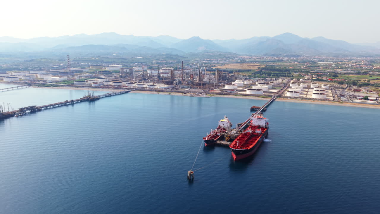 Drone shot focused on two stationary red oil tankers docked at a pier, then tilting up to reveal the refinery, storage tanks, fields, and mountains