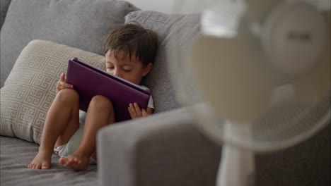 Pequeño-Y-Lindo-Niño-Latino-Con-Un-Mameluco-Azul-Claro-Sentado-En-Un-Sofá-Gris-Disfrutando-De-Una-Película-En-Una-Tableta-Morada