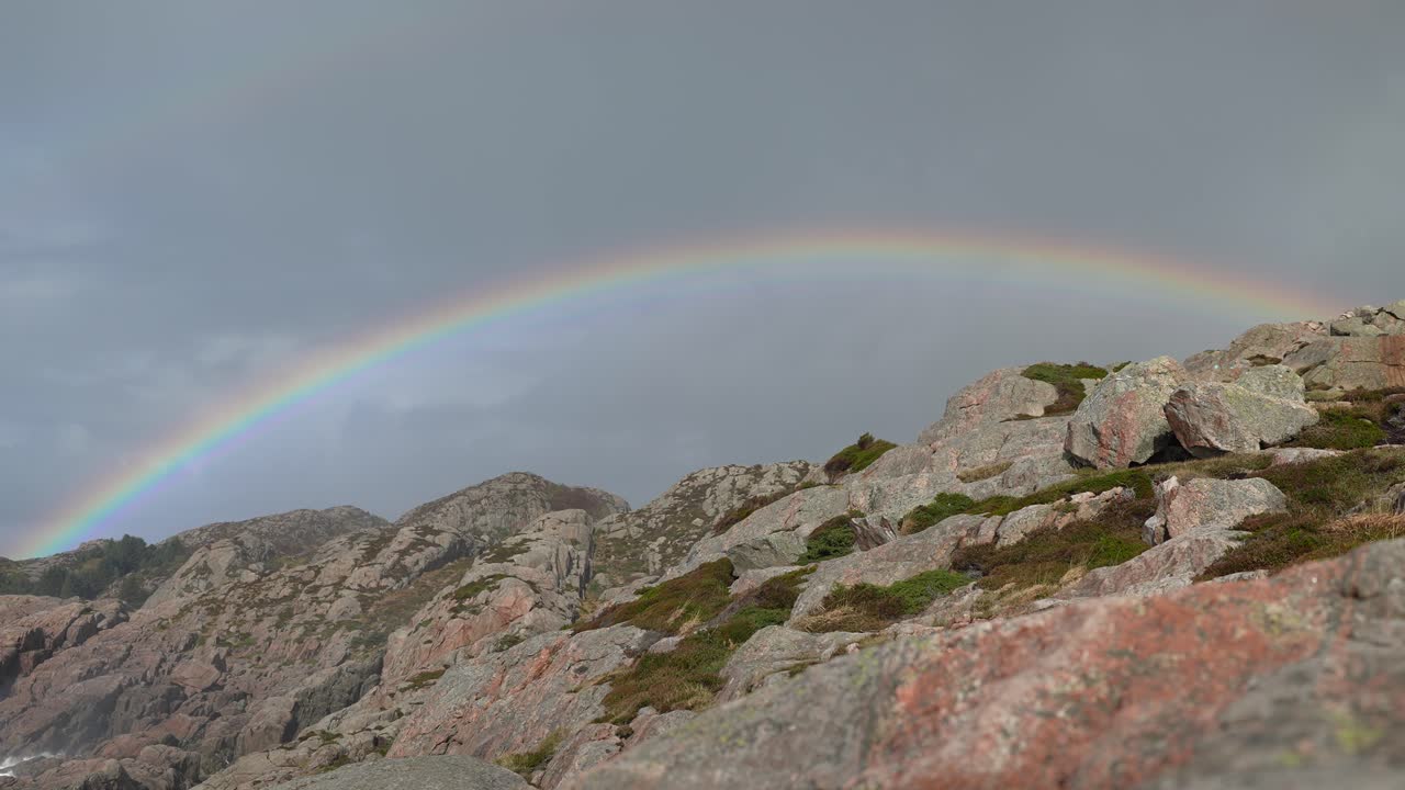 A vibrant rainbow stretches across the sky, creating a stunning contrast against rocky cliffs and patches of green moss under a dramatic sunset sky
