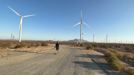 Mann,-Der-Auf-Der-Unbefestigten-Straße-Im-Windmühlenpark-Spaziert,-Drehende-Blätter-Von-Windturbinen-An-Einem-Sonnigen-Tag,-Statischer-Schuss