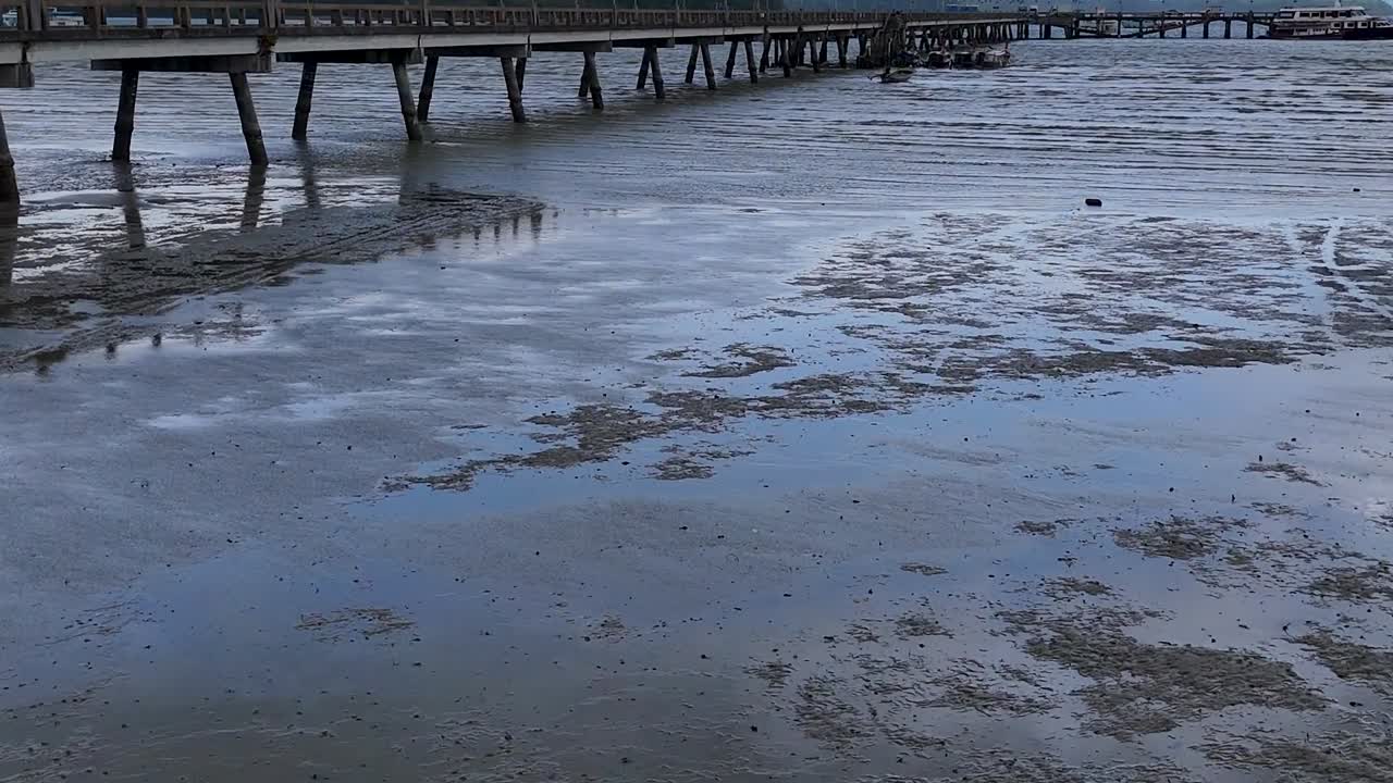 A serene view of wet sand reflecting the sky near a wooden pier during low tide.