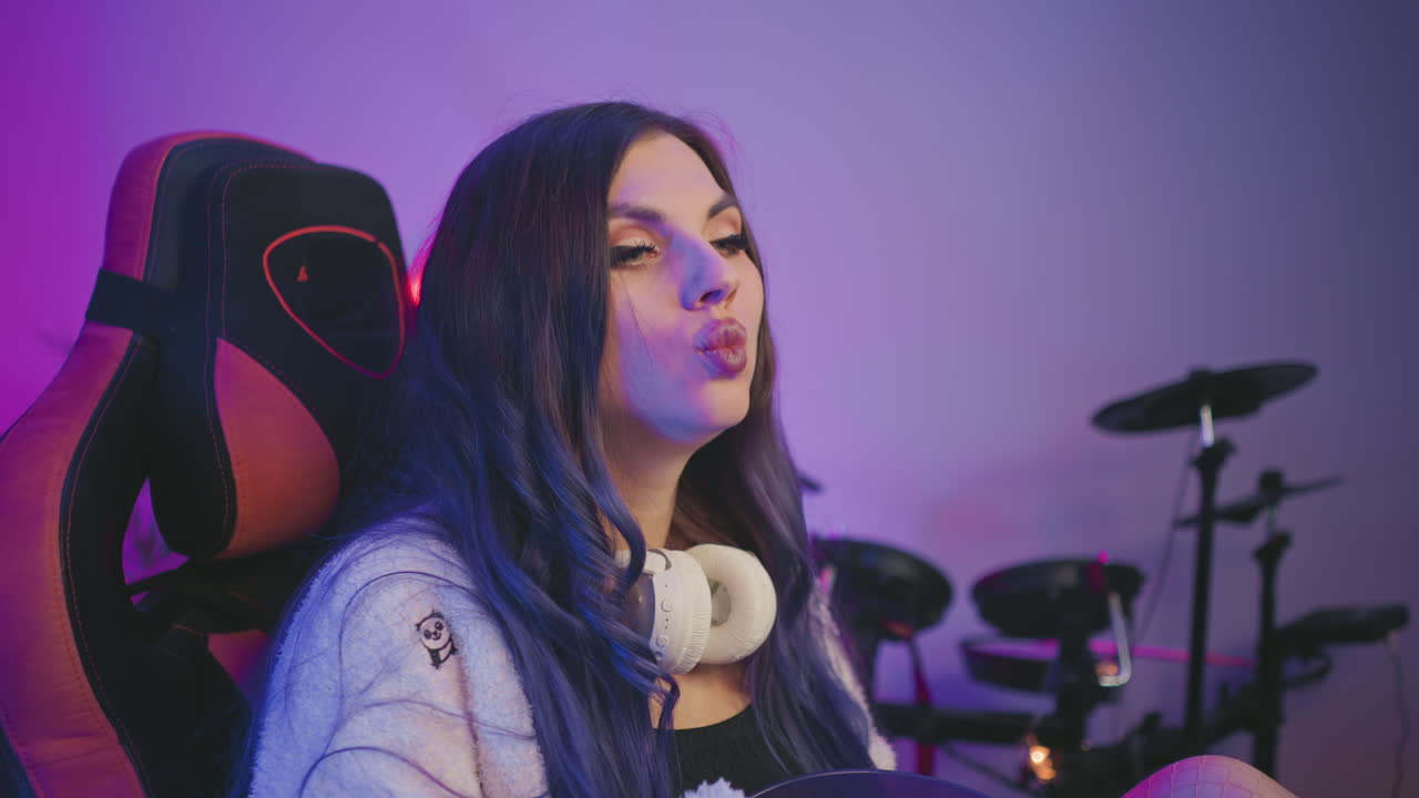 Young lady with painted eyes sits comfortably in gaming chair holding popcorn bowl, staring attentively at something in distance with electronic drum set slightly blurred in background