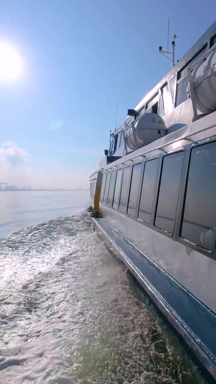 Eine vertikale 9:16-Aufnahme aus der Ich-Perspektive von einer fahrenden Fähre, die die weiße Gischt und eine entfernte Stadtsilhouette einfängt. Ideal für mobile Reise- und maritime Themen in sozialen Medien.