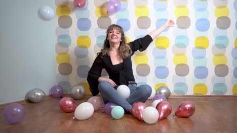 Young-Woman-Sitting-Around-Balloons-and-Throwing-Balloons-to-the-Camera-Against-Colorful-Background