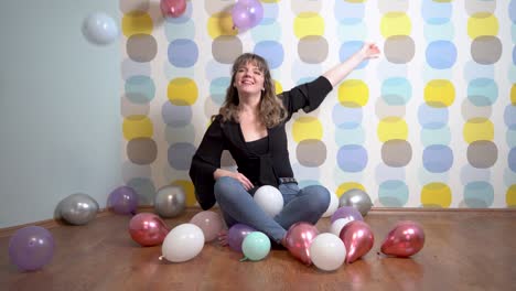 mujer joven sentada alrededor de globos y lanzando globos a la cámara contra un fondo colorido