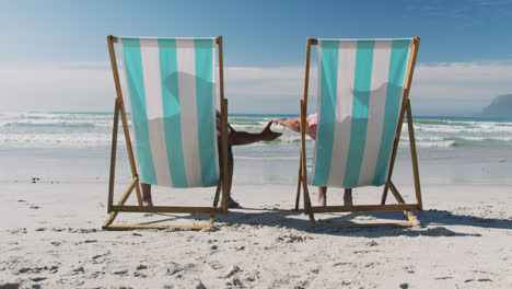 Senior-african-american-couple-sitting-on-sunbeds-and-holding-hands-at-the-beach