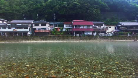 Drone-captures-serene-motion-above-river,-drawing-closer-to-quaint-Japanese-residences-by-the-tranquil-shore