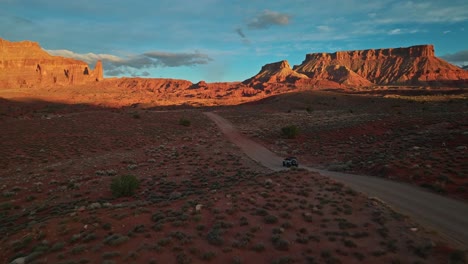 golden sun lights up red cliffs and open road through rugged desert landscape at daybreak as suv traverses dirt path, moab utah usa