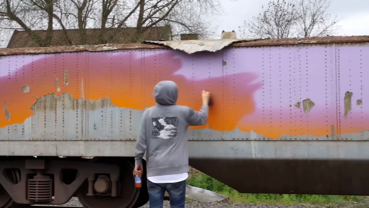 Vista posteriore di una figura incappucciata che spruzza colore sul lato di un vagone merci usurato, con il braccio alzato verso un pannello rivettato mentre una bomboletta di riserva riposa nell'altra mano, posizionata accanto a un binario di ghiaia in un aspro cortile industriale.