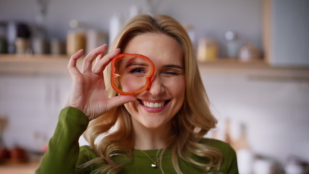 Funny woman holding pepper slice peeping through at bright kitchen portrait