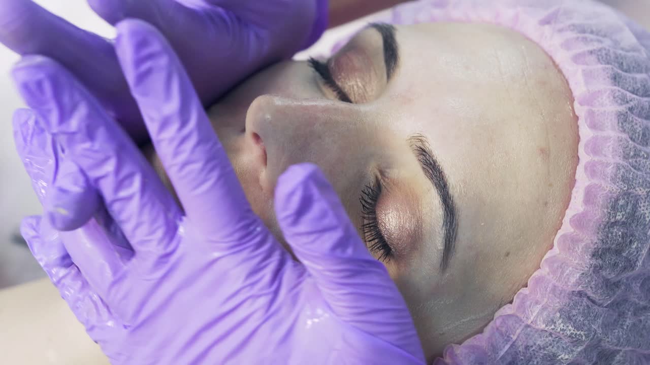 Relaxed woman in medical cap is having face massage at spa salon. Cosmetologist in medical purple gloves is touching girl's face.