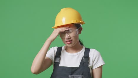 녹색 스크린 배경 스튜디오에 서 있는 동안 두통을 가진 보호 안경과 안전 헬을 착용 한 아시아 여성 노동자의 근접