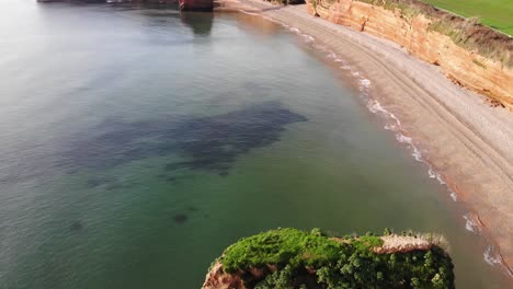 aerial flying over sea stack with green vegetation with view of ladram bay beach