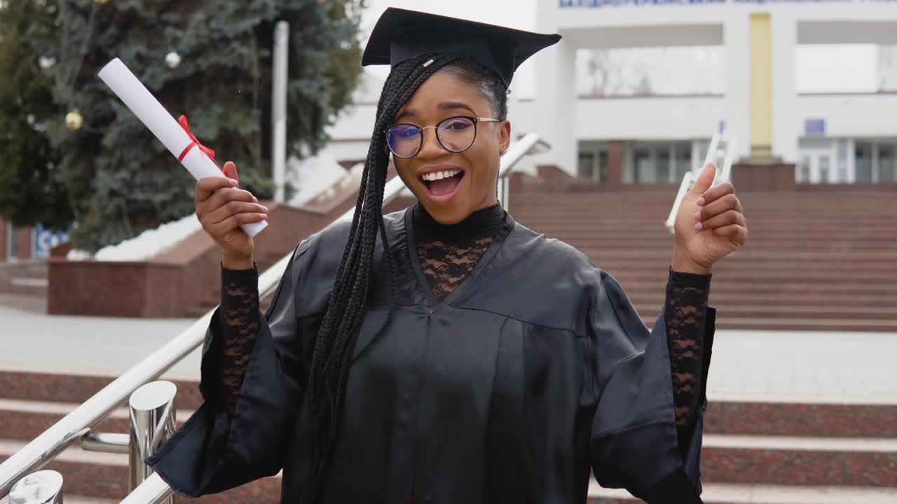una mujer joven en la universidad en el manto del maestro sostiene un diploma mientras baila felizmente y mira a la cámara. retrato frente a la entrada central de la universidad en segundo plano