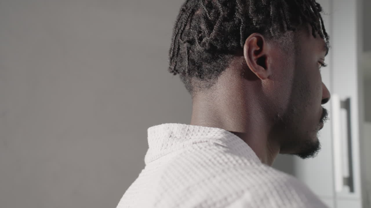 Close-up of man in bathrobe holding spoon and preparing to stir a drink in clear glass cup. Modern kettle and minimalist kitchen design visible in background, soft morning light enhancing the scene