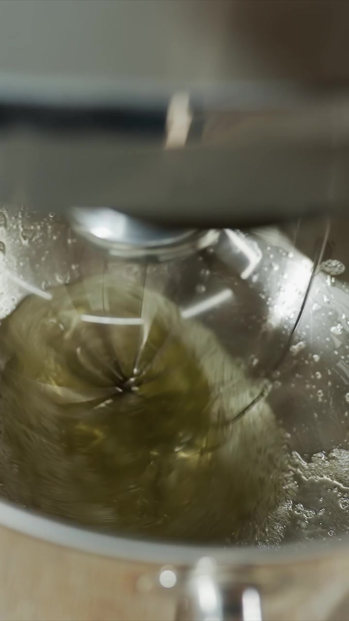 Primo piano di un liquido verde mescolato da una frusta metallica. La miscela si agita continuamente mentre si amalgama uniformemente. Un piano di lavoro in legno è visibile brevemente, indicando un ambiente cucina interno