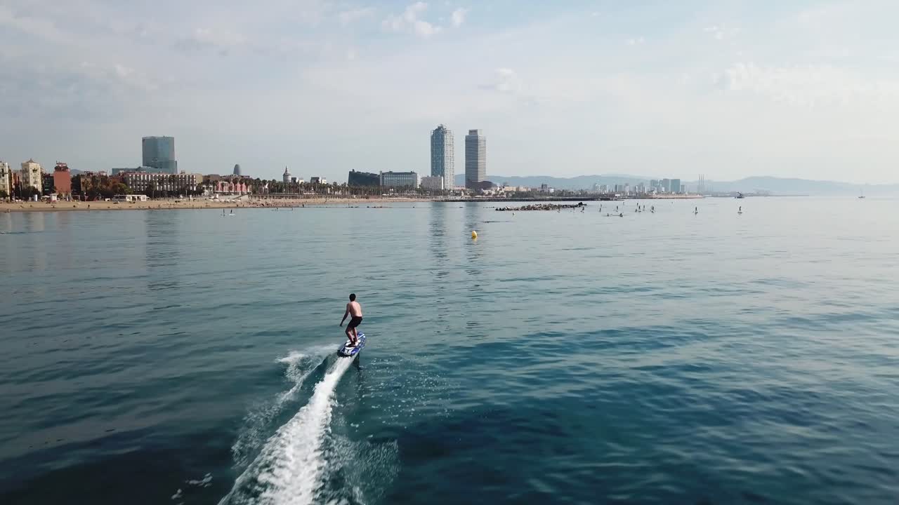 toma aérea de un surfista rápido montando una tabla de surf eléctrica frente al icónico horizonte de barcelona en un día soleado