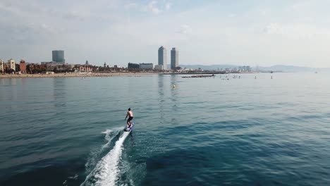 Luftaufnahme-Eines-Schnellen-Surfers,-Der-An-Einem-Sonnigen-Tag-Vor-Der-Berühmten-Skyline-Von-Barcelona-Auf-Einem-Elektrischen-Surfbrett-Reitet