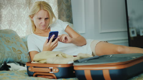 a woman uses a smartphone lies next to an open suitcase with clothes