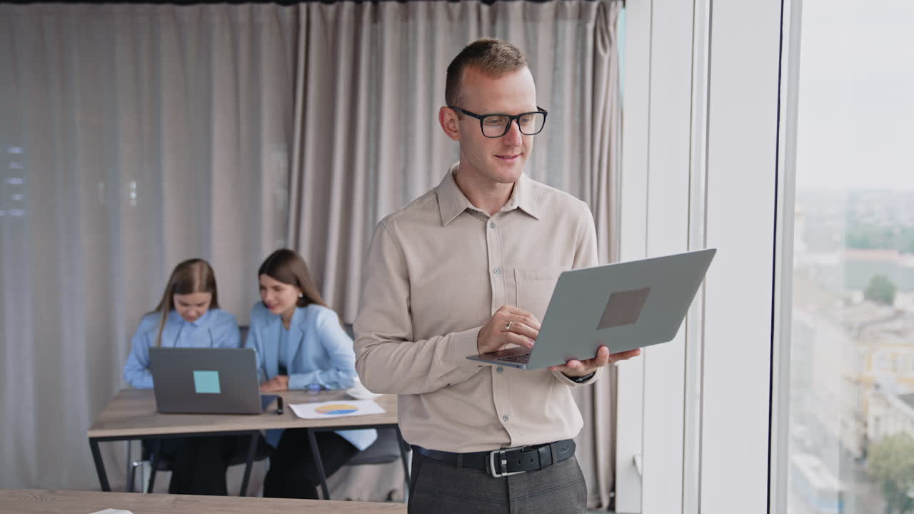 Male employee wearing glasses holds a laptop standing at the window. Female colleagues sitting at desk in front of laptop at backdrop.