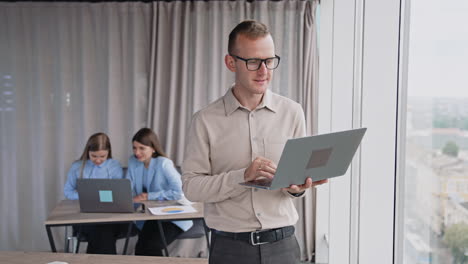 male employee wearing glasses holds a laptop standing at the window. female colleagues sitting at desk in front of laptop at backdrop.