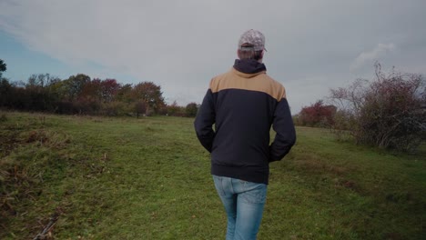 tall scandinavian guy with cap and jacket explores the fields of vårhallarna in autumn, simrishamn sweden österlen - tracking medium shot from behind