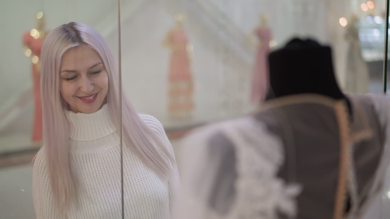 fashion lover smiles while gazing at white lace gown behind glass display in mall boutique, hands resting on glass as soft lighting highlights delicate lace and mannequins in background