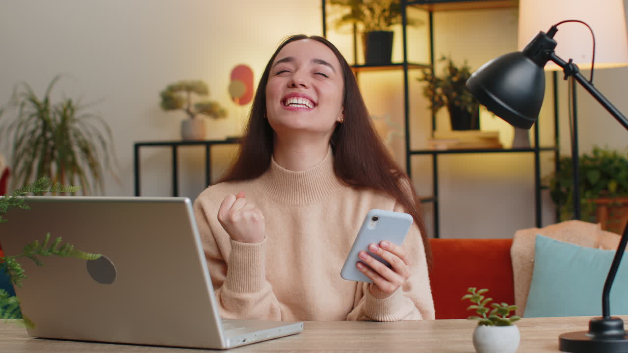 mujer caucásica en la mesa de casa usar teléfono inteligente celebrar el éxito victoria ganar jugar juego en línea