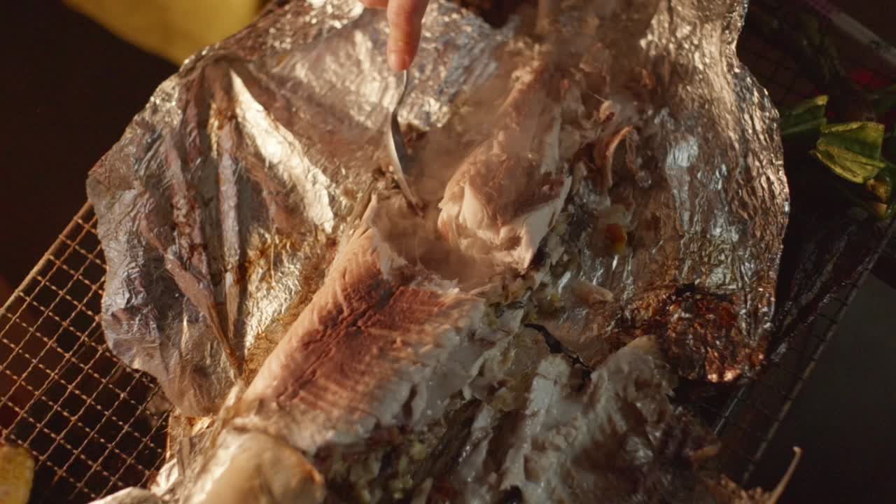 Freshly barbecue baked fish on foil broken in half using spoon by hand, filmed as closeup in slow motion style