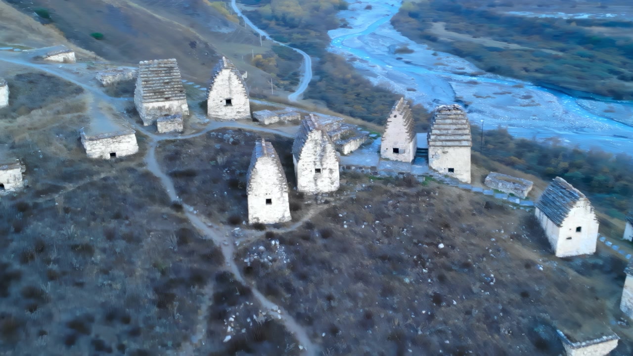 Yumuşak gün ışığında, çorak ve kayalık bir yamaçta dağınık halde duran küçük, yıpranmış taş kulübelerden oluşan terk edilmiş bir kırsal yerleşimin drone görüntüsü.