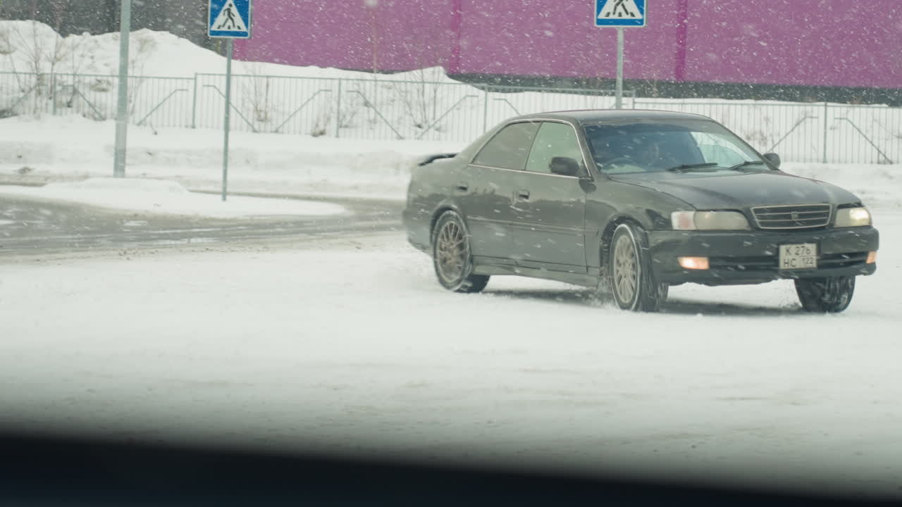 car maneuvers in zigzag motion on snow-covered road during active snowfall with snowflakes visibly swirling through frame background includes pedestrian sign, fence, and industrial building
