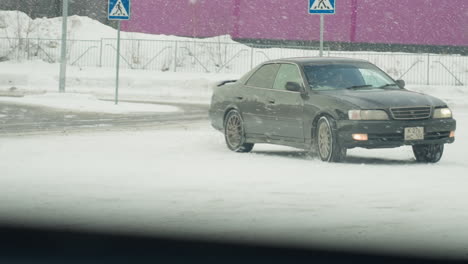 car maneuvers in zigzag motion on snow-covered road during active snowfall with snowflakes visibly swirling through frame background includes pedestrian sign, fence, and industrial building
