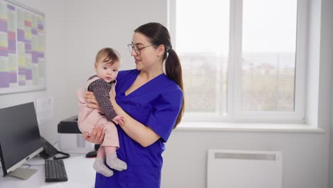 a happy brunette girl in a blue uniform a doctor in glasses holds a little baby girl in her arms and finds a common language with her during prevention in a modern clinic with a pediatrician