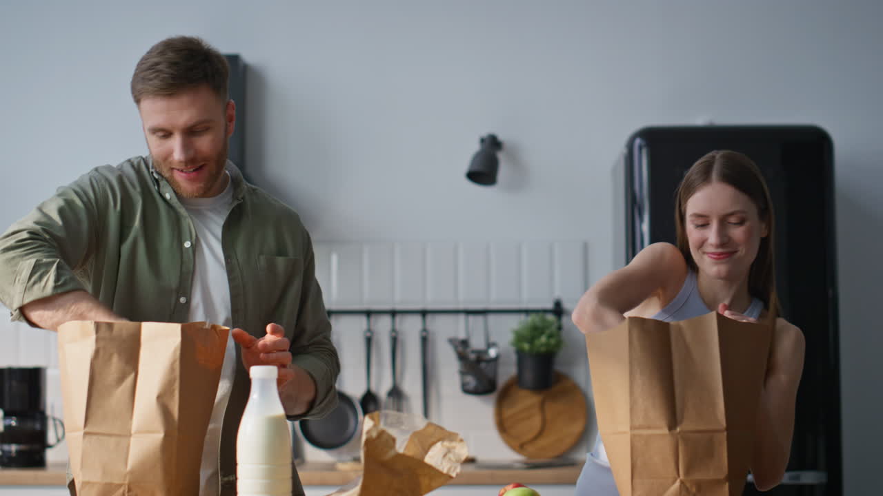 Laughing spouses unpacking groceries in home kitchen fooling together closeup