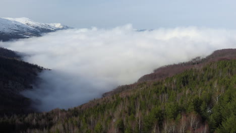 Vista-Aérea-De-Las-Hermosas-Laderas-Del-Valle-De-Montaña-Cubiertas-De-Nubes-Flotantes-Pico-De-Montaña-Cubierto-De-Nieve-A-Lo-Lejos-El-Día