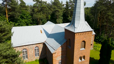 aerial drone ascending and flying higher in front of a old red colored historic brick church in alatskivi during a summer sunny day, a large pine tree and its branches are in the foreground, blue sky