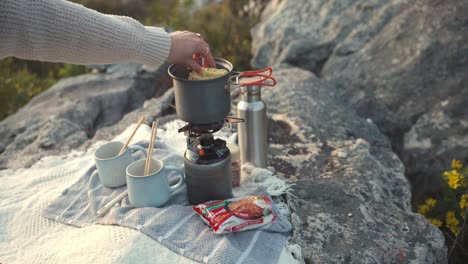 anonymous woman drops raw noodles into boiling saucepan on portable stove at campsite in england, uk