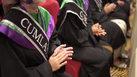 close up happy graduates in hats and robes siting in row and applauding