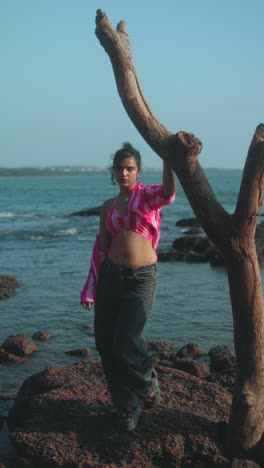 young woman standing near tree by the beach, wearing a pink shirt and jeans