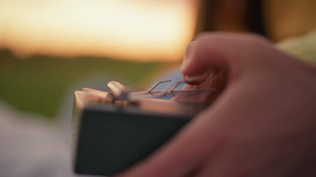 closeup hands playing kalimba during sunset, soft bokeh background and warm golden light, thumb plucking metal tines on wooden box, calm evening picnic scene, solo musician composing gentle melody
