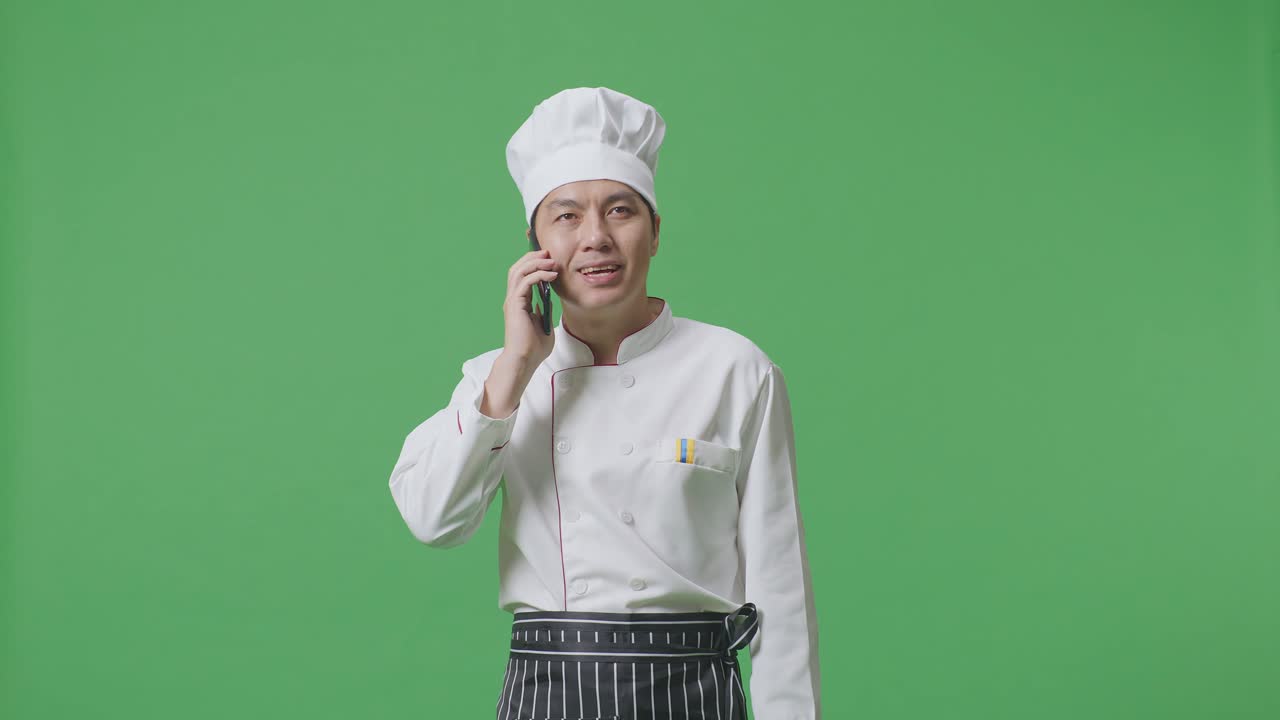 Asian Man Chef Talking On Smartphone And Looking Around While Standing In The Green Screen Background Studio