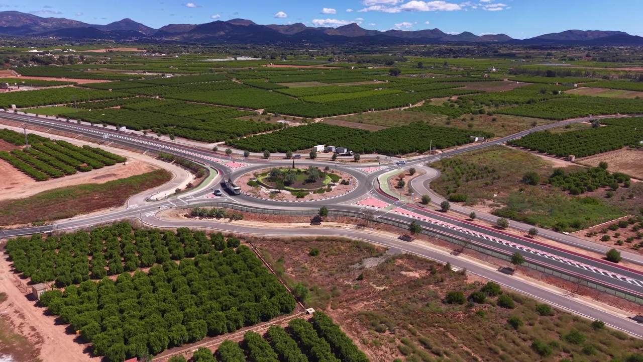 Gran intersección de rotonda vial rodeada de campos agrícolas verdes y huertos en la España rural, con una cordillera visible bajo un cielo azul.