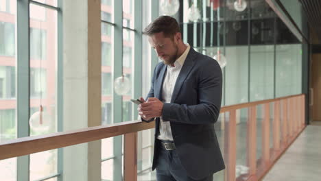 Un-Joven-Y-Atractivo-Hombre-De-Negocios-Barbudo-Con-Una-Chaqueta-Azul-Consulta-Atentamente-Su-Teléfono-Inteligente-Y-Escribe-En-él-1