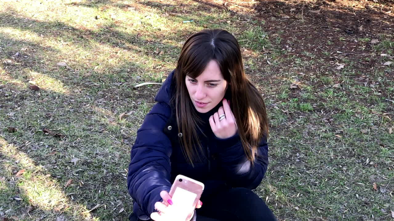 cámara lenta de una joven tomando selfies con un smartphone usando una cámara frontal sentada en el suelo en un parque urbano