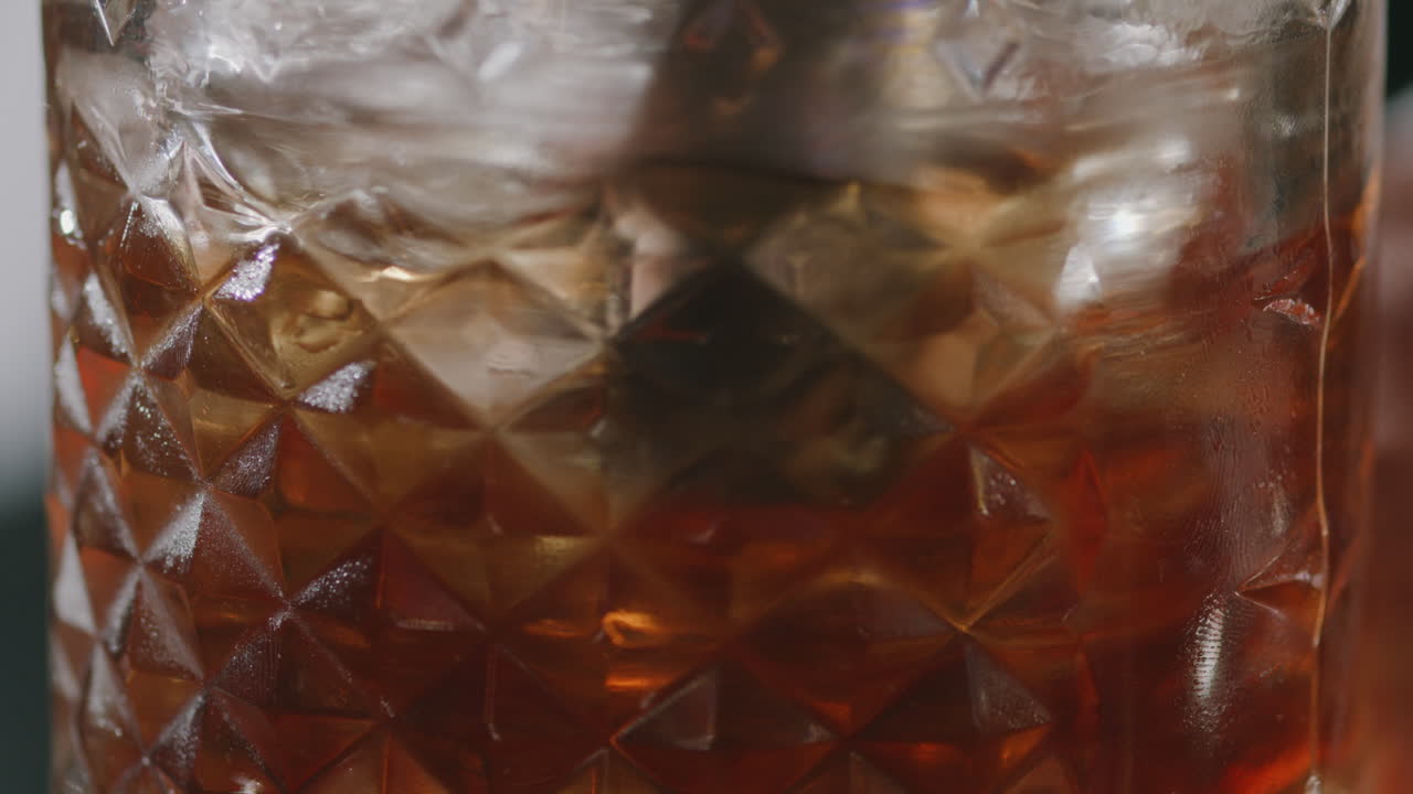 Close-up of a whiskey glass with ice being stirred with a bar spoon. The movement highlights the shine of the liquid and the detail of the ice cubes.