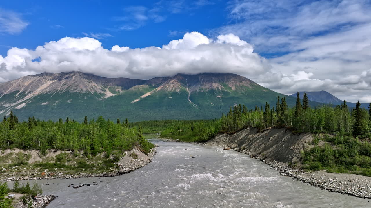 Fiume impetuoso sotto la catena montuosa dell'Alaska. Acqua grigia e veloce che scava un canyon boschivo ai piedi delle cime dell'Alaska avvolte dalle nuvole