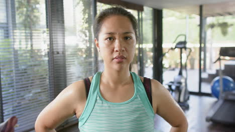 serious asian woman with arms akimbo looking at camera in sunny home gym, slow motion