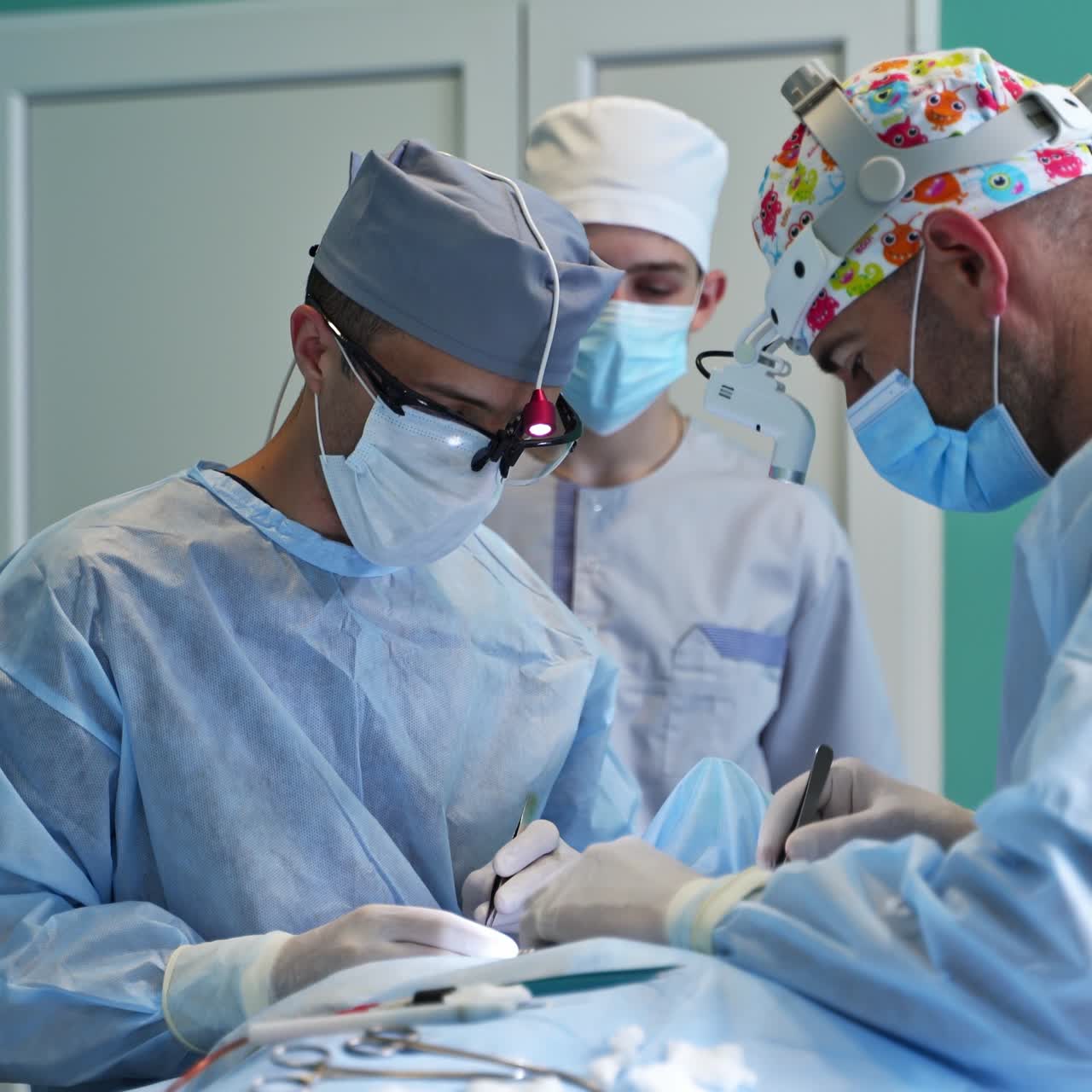 Læger bruger metalinstrumenter (forceps) under en operation. En studerende overværer operationen i baggrunden.